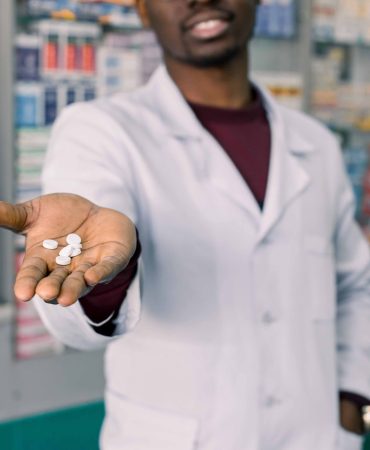 African-american young doctor or pharmacist offering pill in hand, standing over pharmacy background.